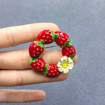 Red strawberry-shaped brooch held between fingers against a gray background