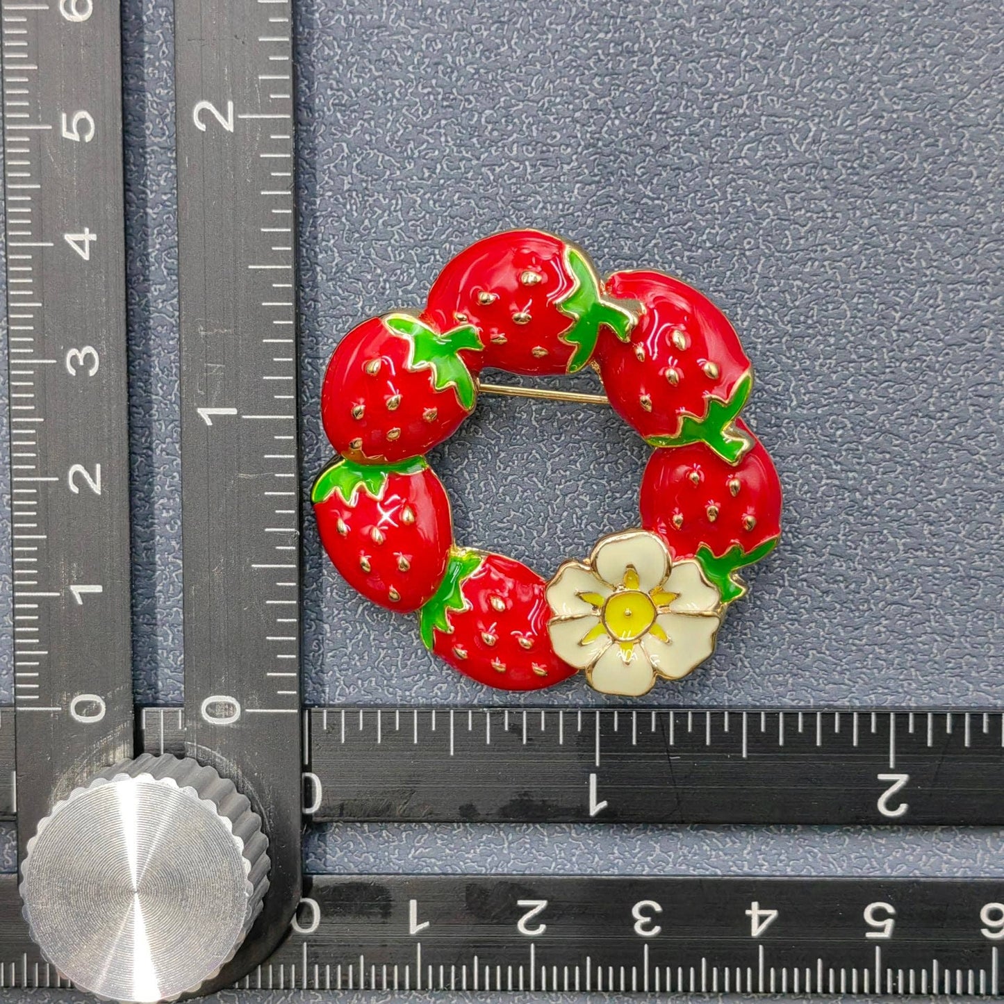 Strawberry wreath brooch next to a ruler on a gray background