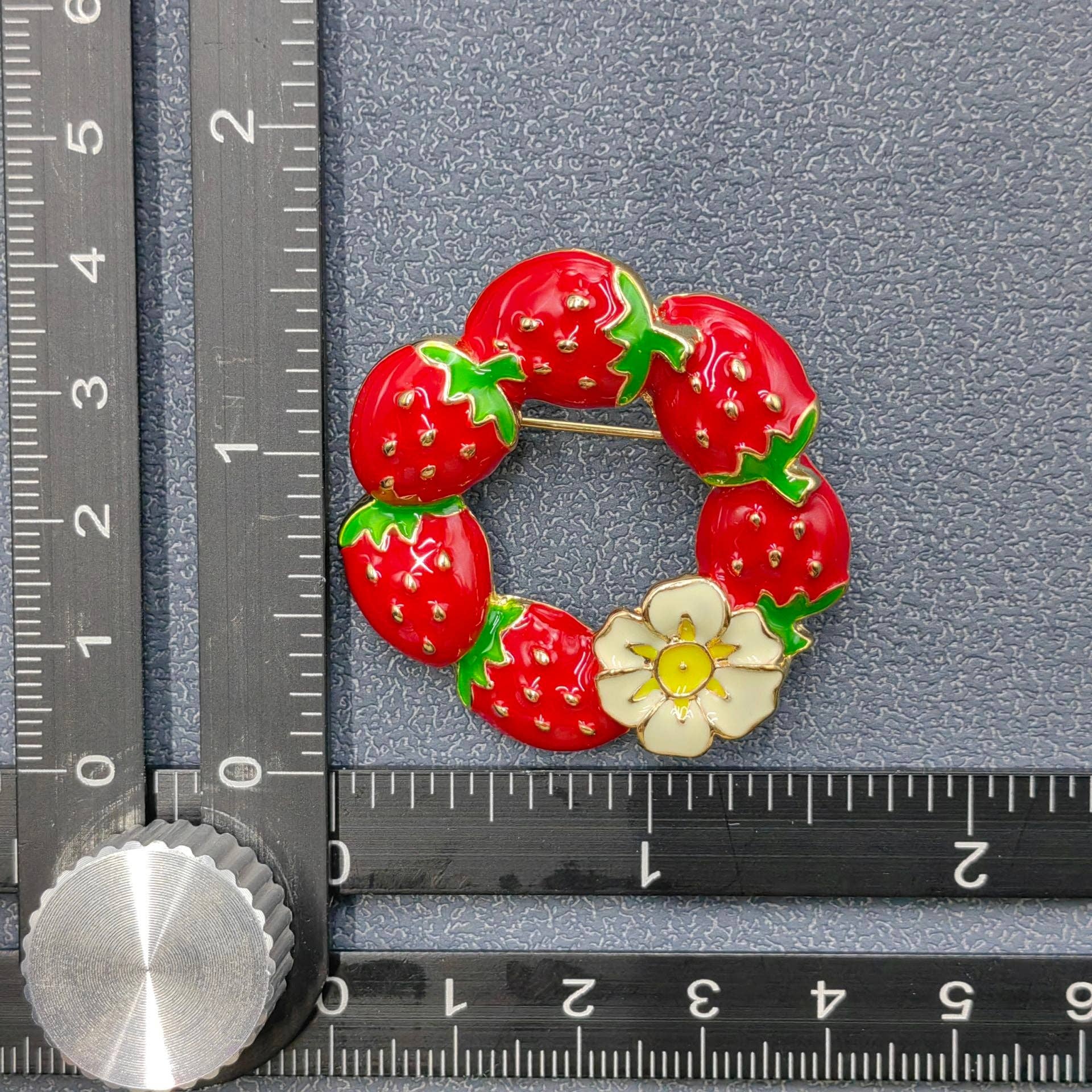 Strawberry wreath brooch next to a ruler on a gray background