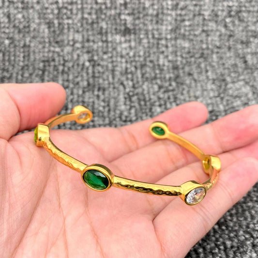 a hand holding a gold adjustable bangle with inlaid green and clear crystal stones