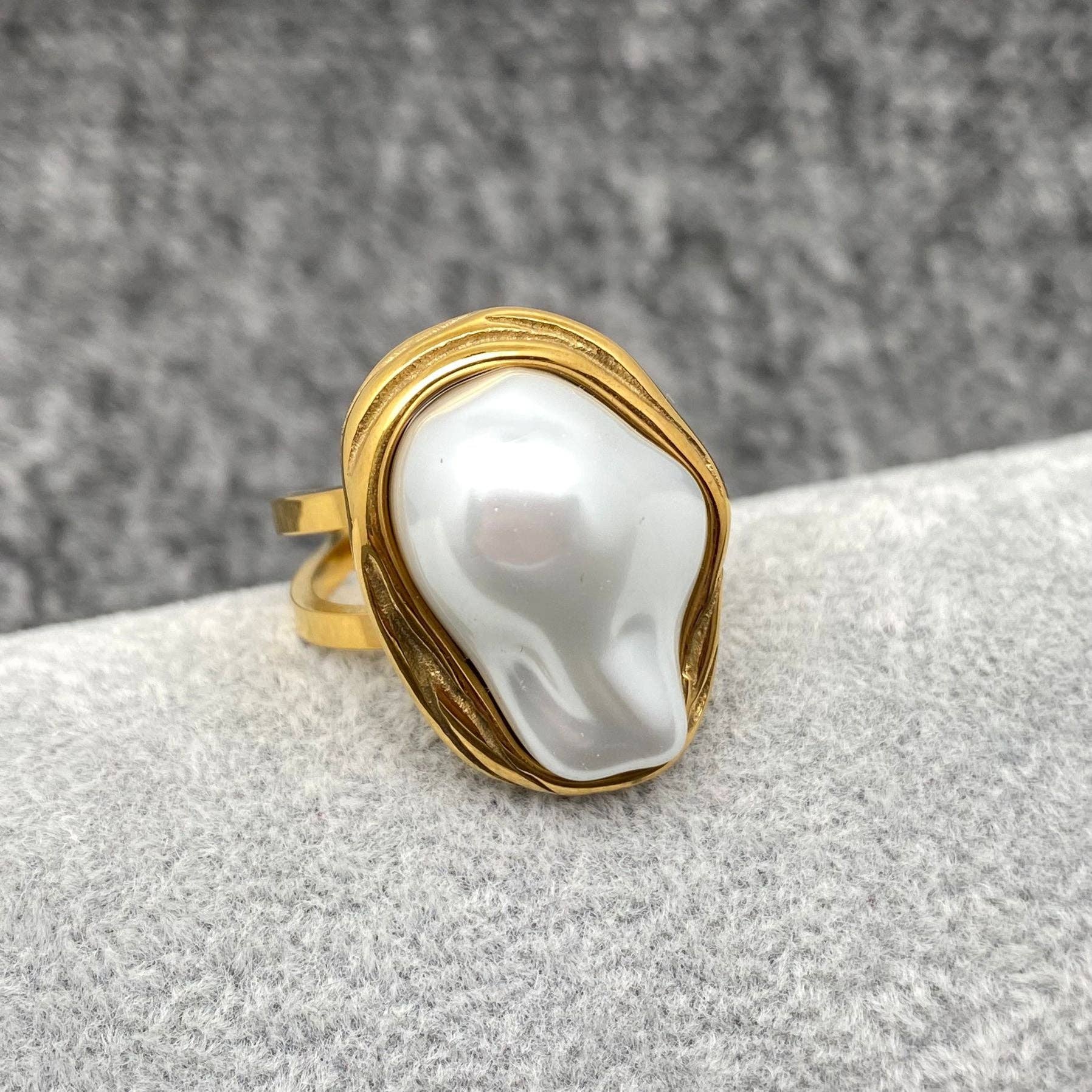 a gold adjustable ring with white baroque style pearl laying on a grey background