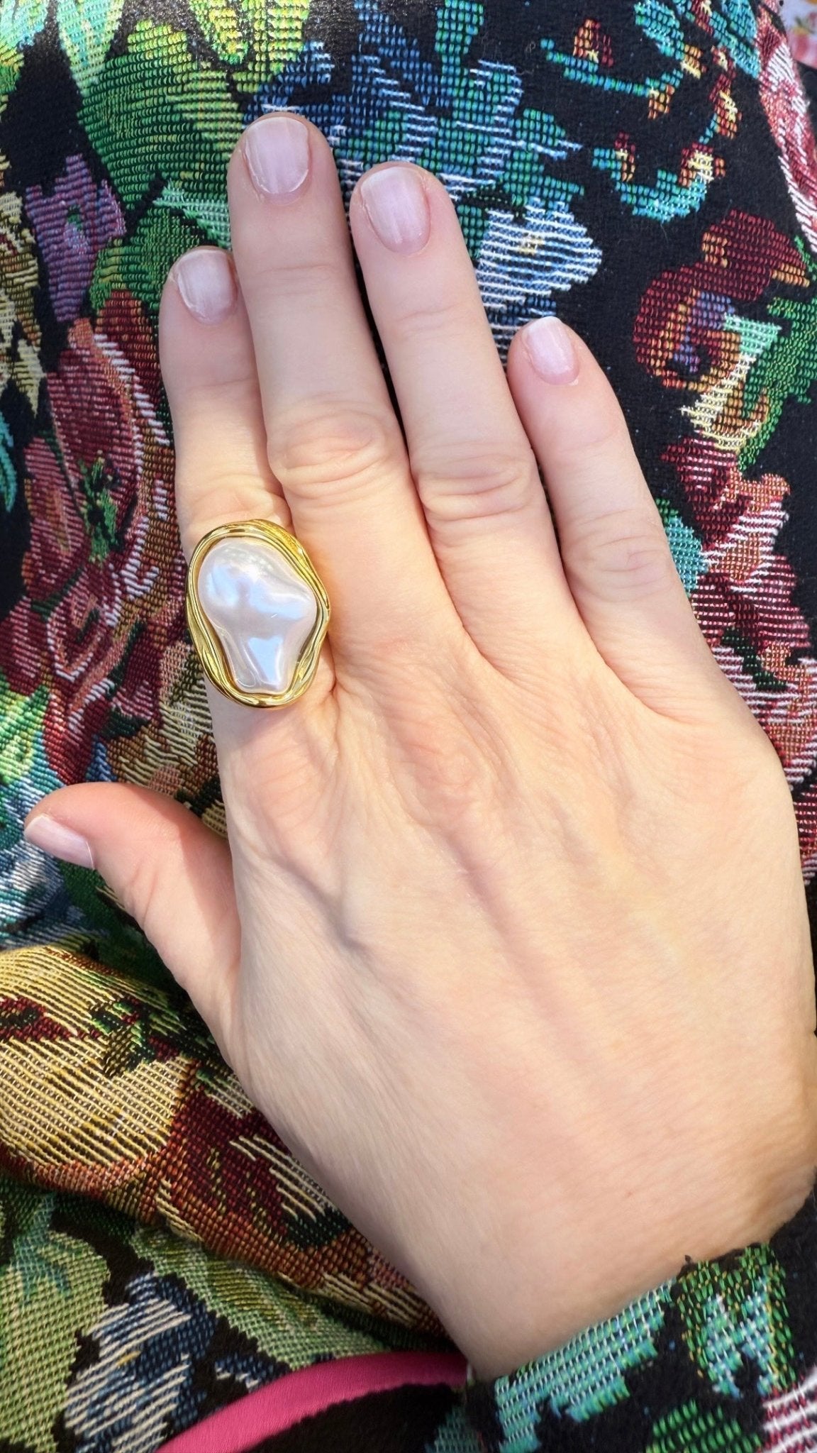 Hand wearing a gold ring with a large baroque pearl on a floral patterned fabric background
