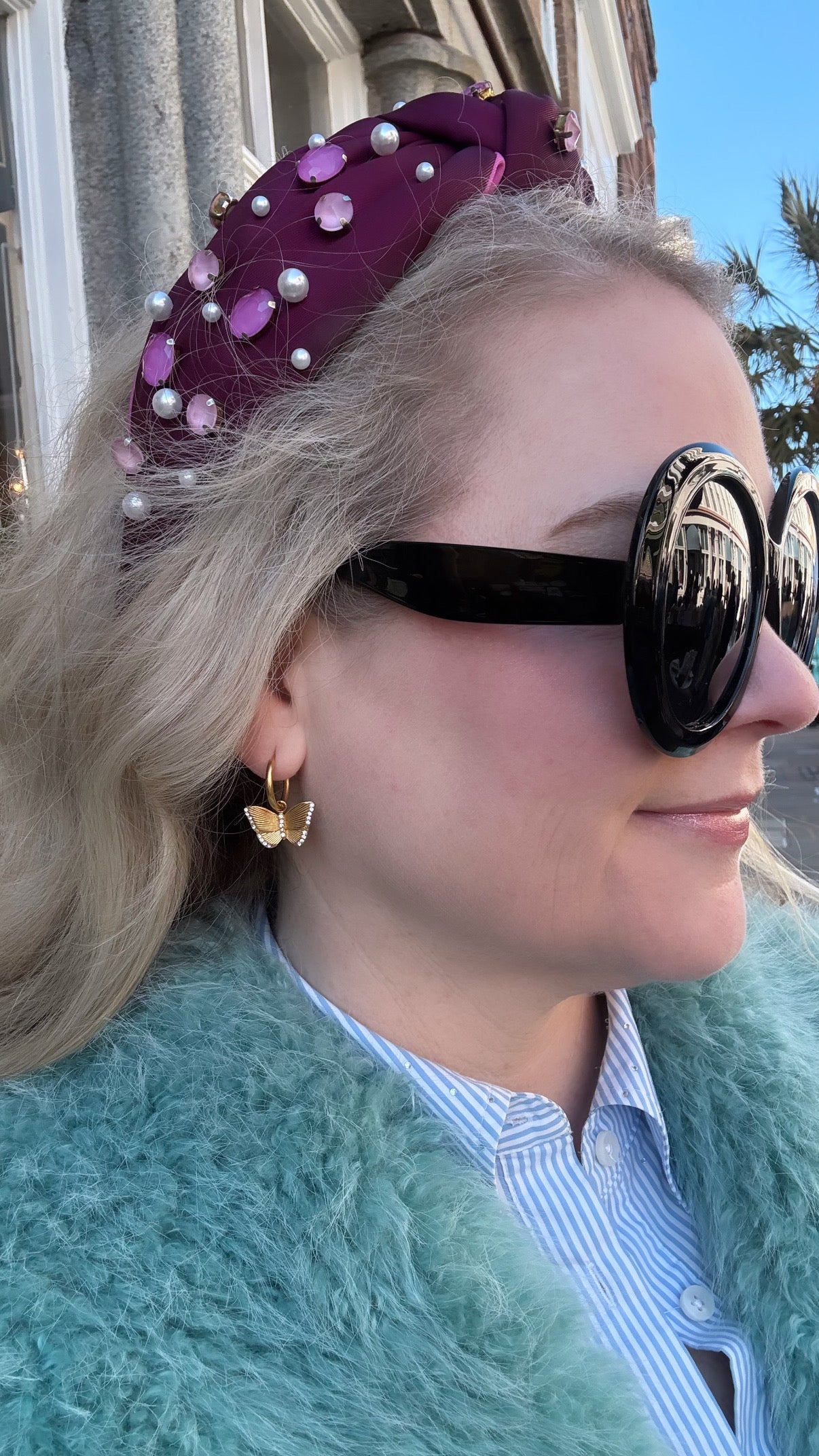 Woman wearing a purple headband with pearls, sunglasses, butterfly and crystal dangle earrings, and a green fur coat.