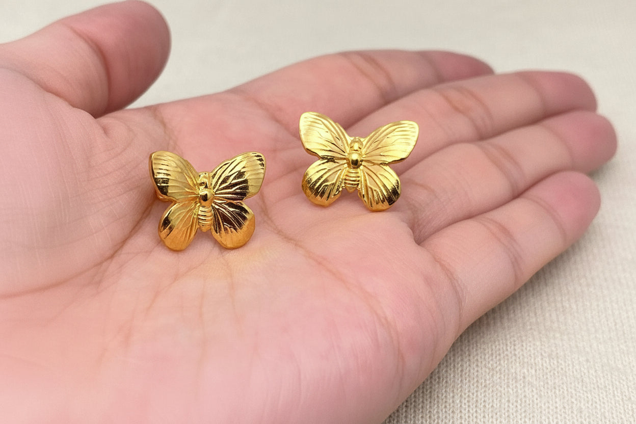 hand holding a pair of gold butterfly stud earrings against an ivory fabric background