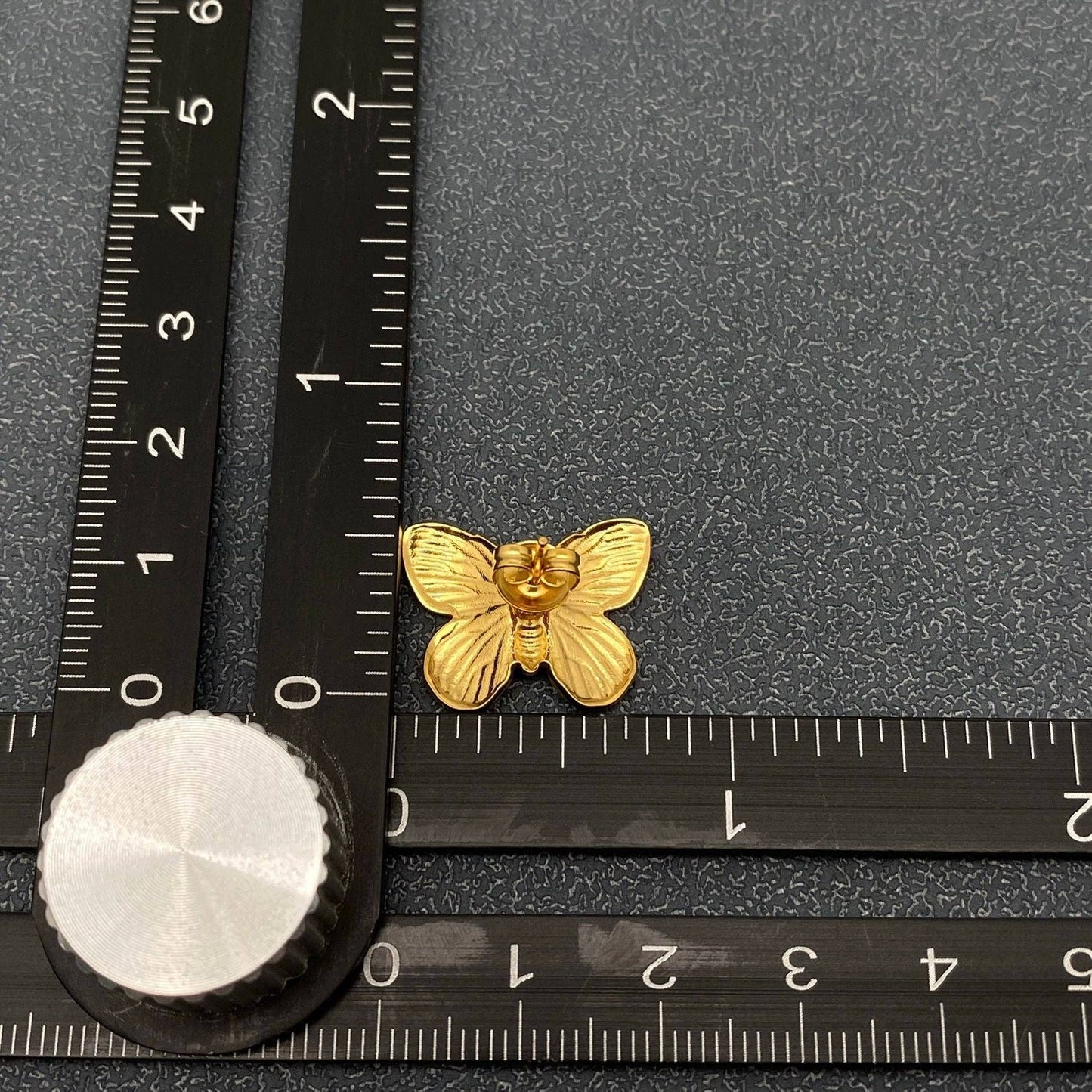 back view of gold butterfly stud earrings next to a ruler for scale against a grey background