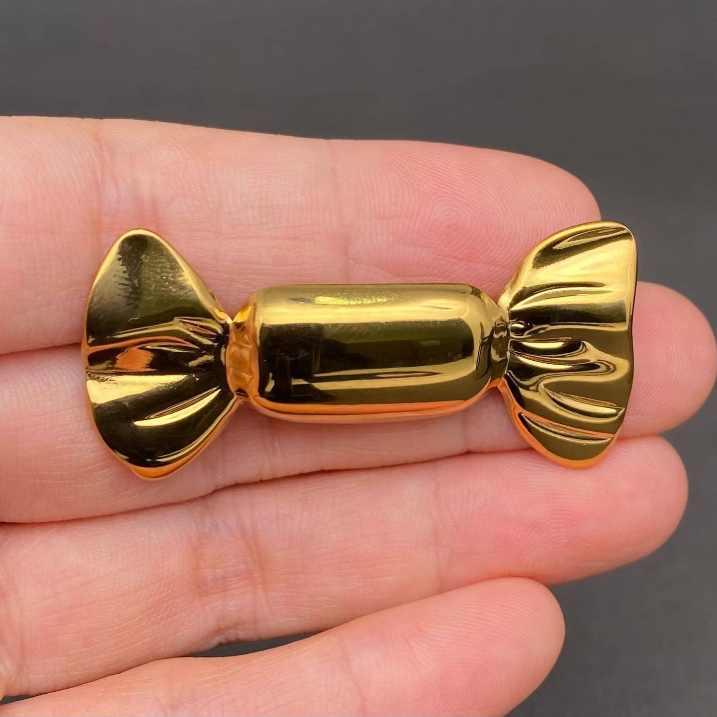 Gold candy wrapper-shaped brooch held between fingers against a dark background