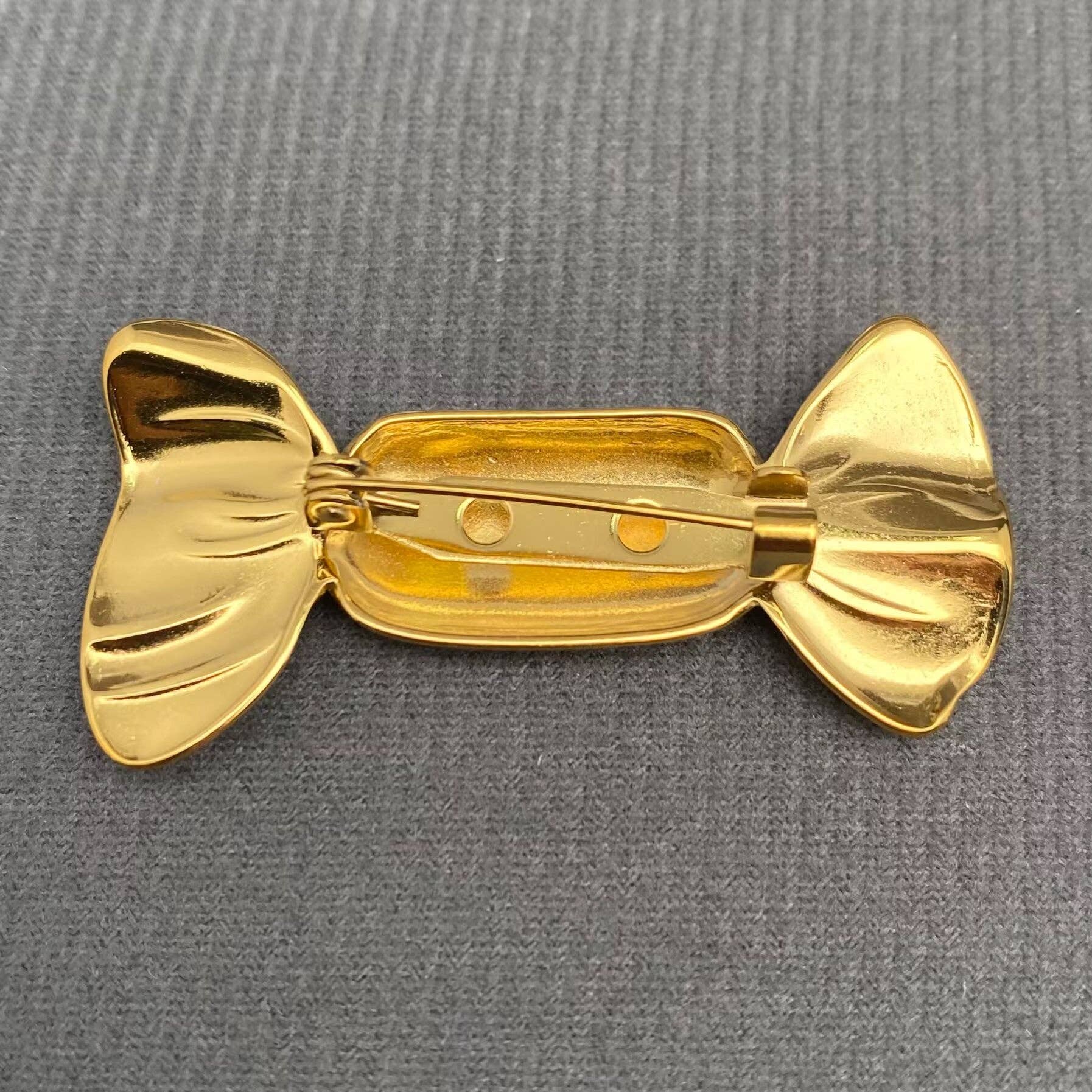 Back view of gold candy wrapper-shaped brooch showing pin closure on a gray textured background