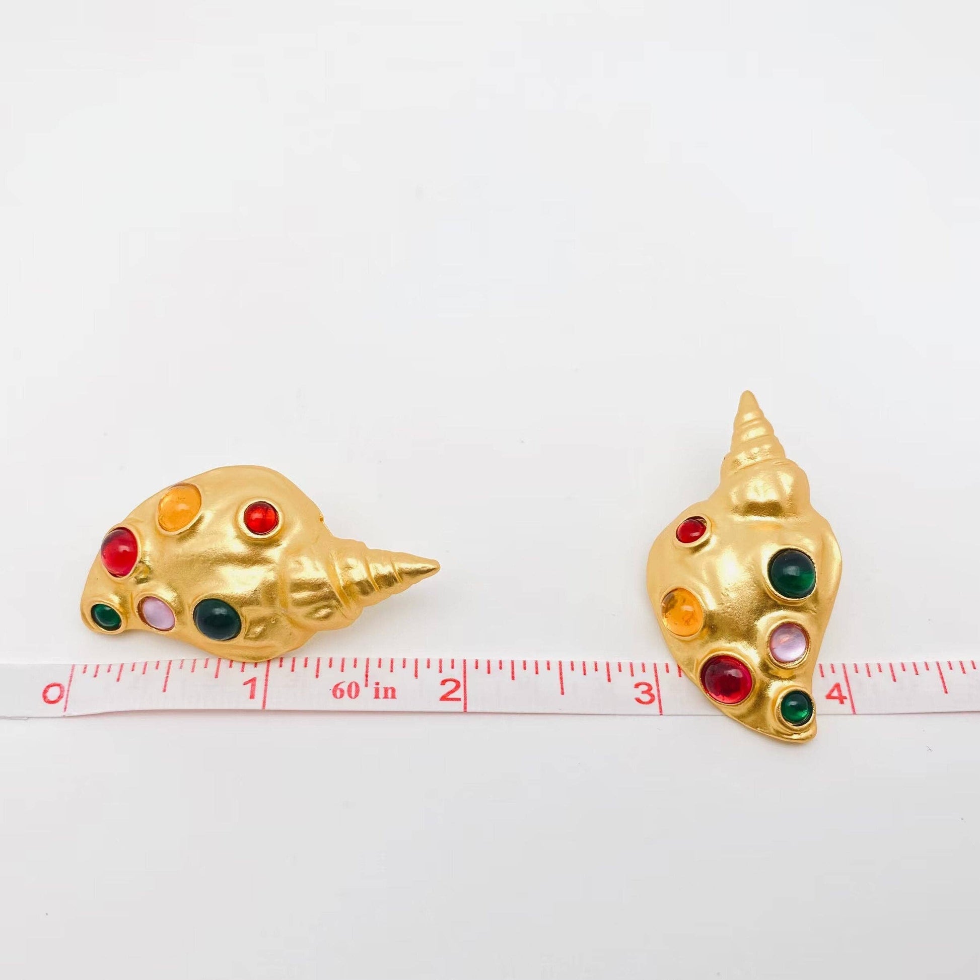 a pair of gold conch earrings with inlaid multi-colored glass stones against a ruler