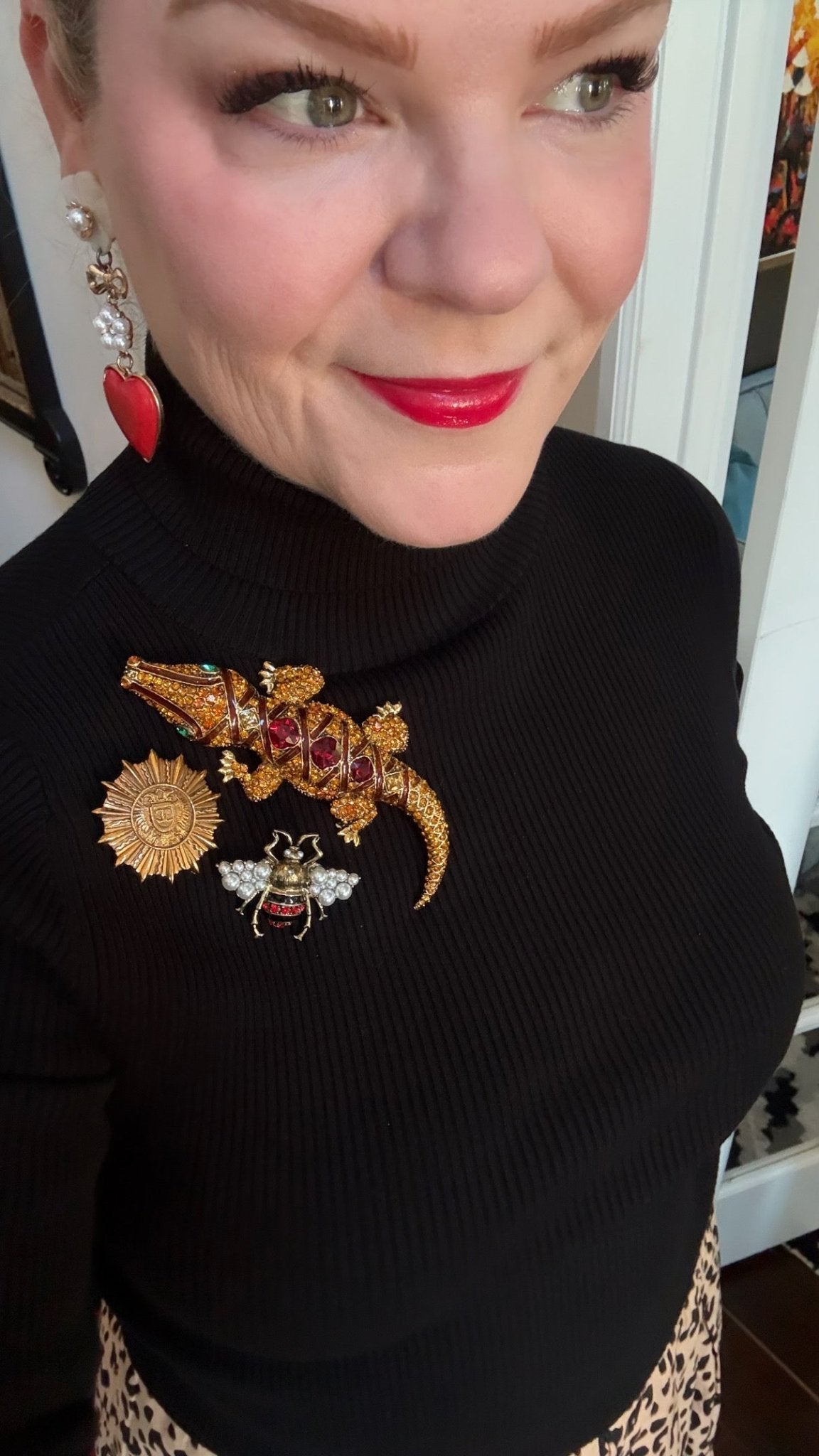 Woman wearing a black sweater with 3 decorative brooches including a bee, sunburst, and alligator and red lipstick.