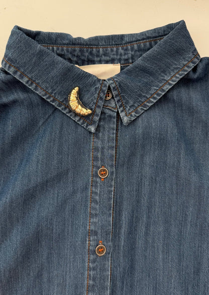 Close-up of a blue denim shirt with a gold croissant brooch on the collar.