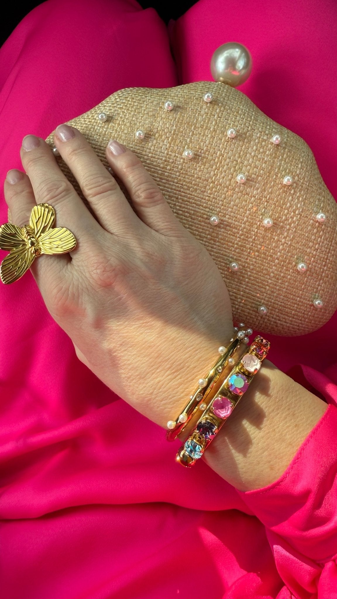 Hand wearing gold butterfly ring and colorful bangles touching a raffia and pearl encrusted clutch on pink fabric background
