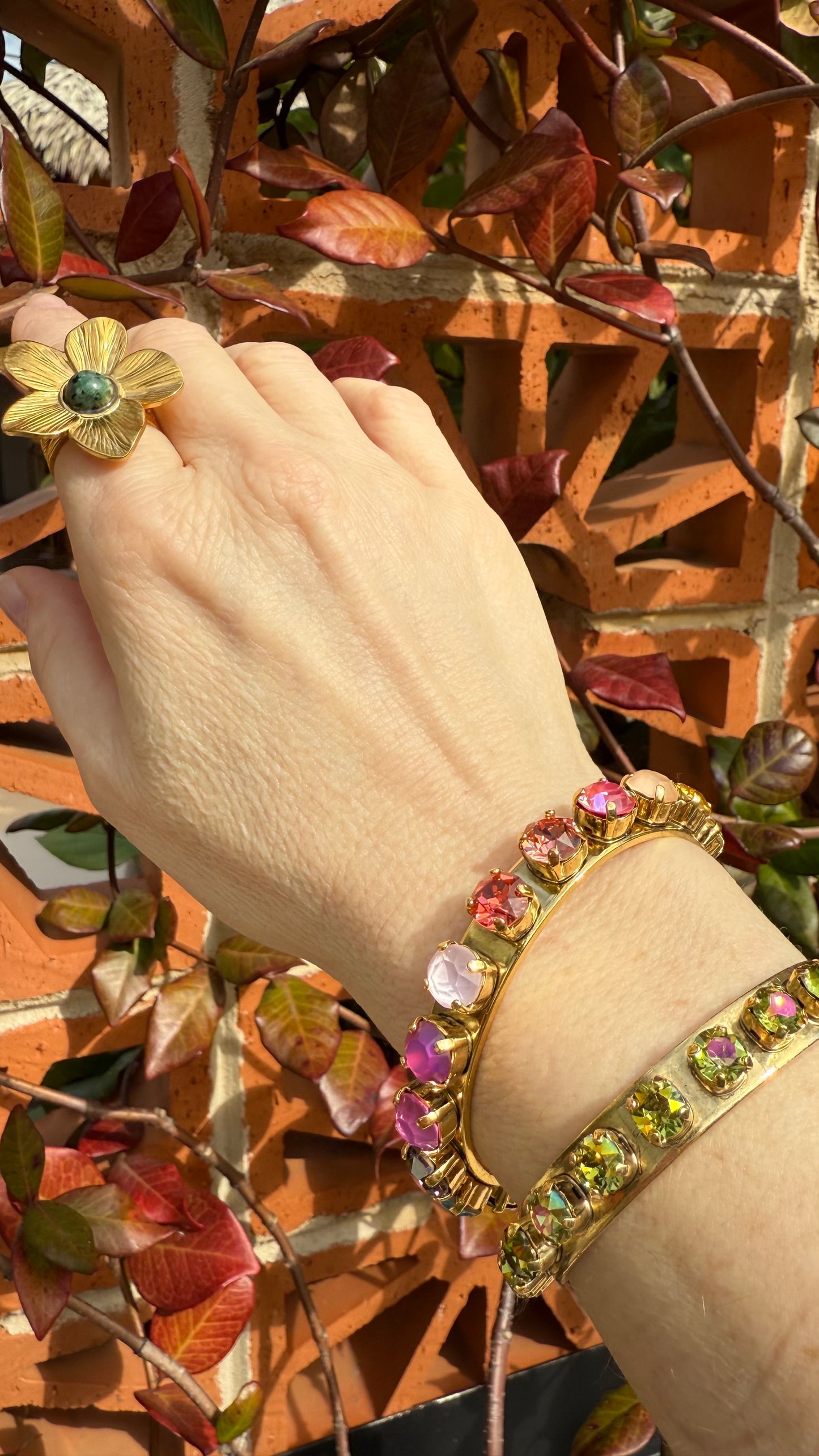 Hand wearing a gold ring with a flower design and a colorful crystal bracelets, set against a leafy textured wall.