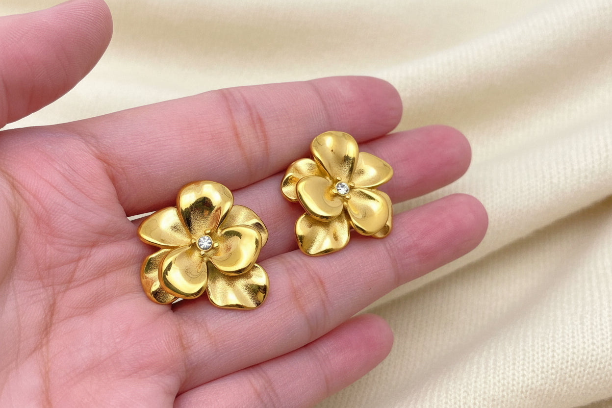Gold flower-shaped earrings held in a hand against an ivory fabric background