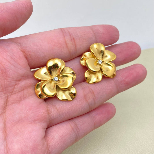 Gold flower-shaped earrings held in a hand against a light background