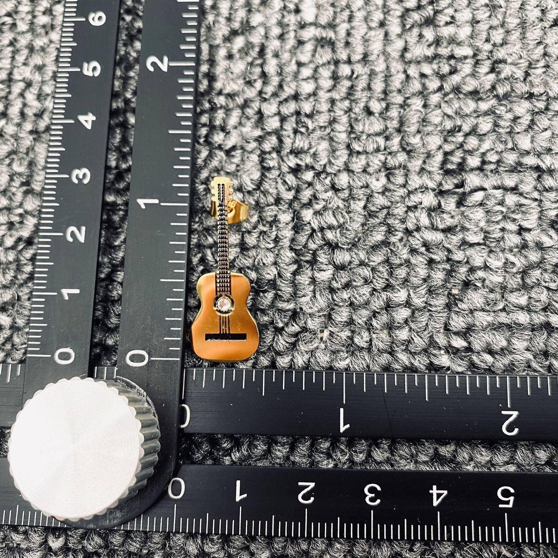Gold guitar shaped earring on a grey textured surface with a ruler for scale.