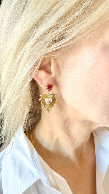 Close-up of a person wearing a gold heart-shaped earring with a red crystal at the top.