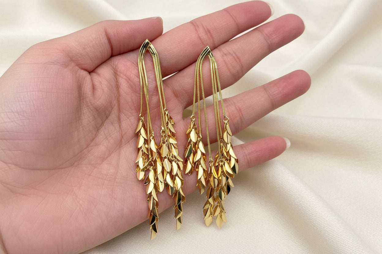 Gold leaf-shaped dangle earrings held in a hand on an ivory fabric background