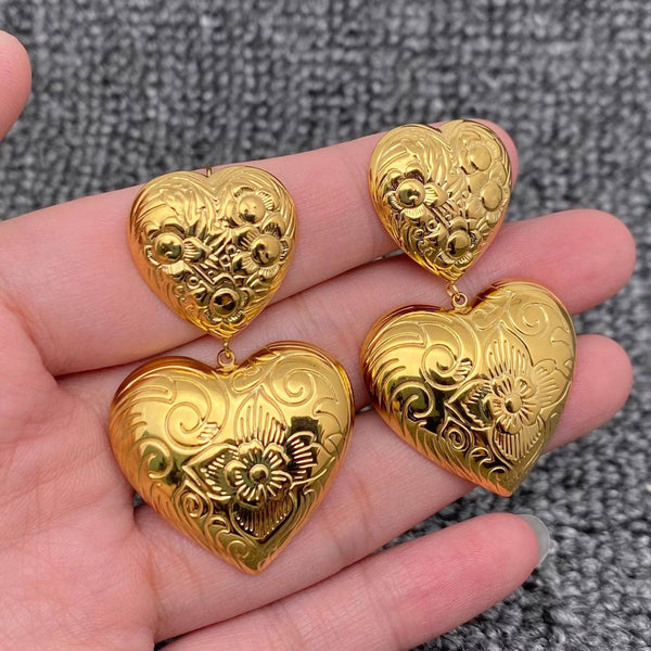 Gold heart dangle earrings with floral engraving held in hand showing two-tier design