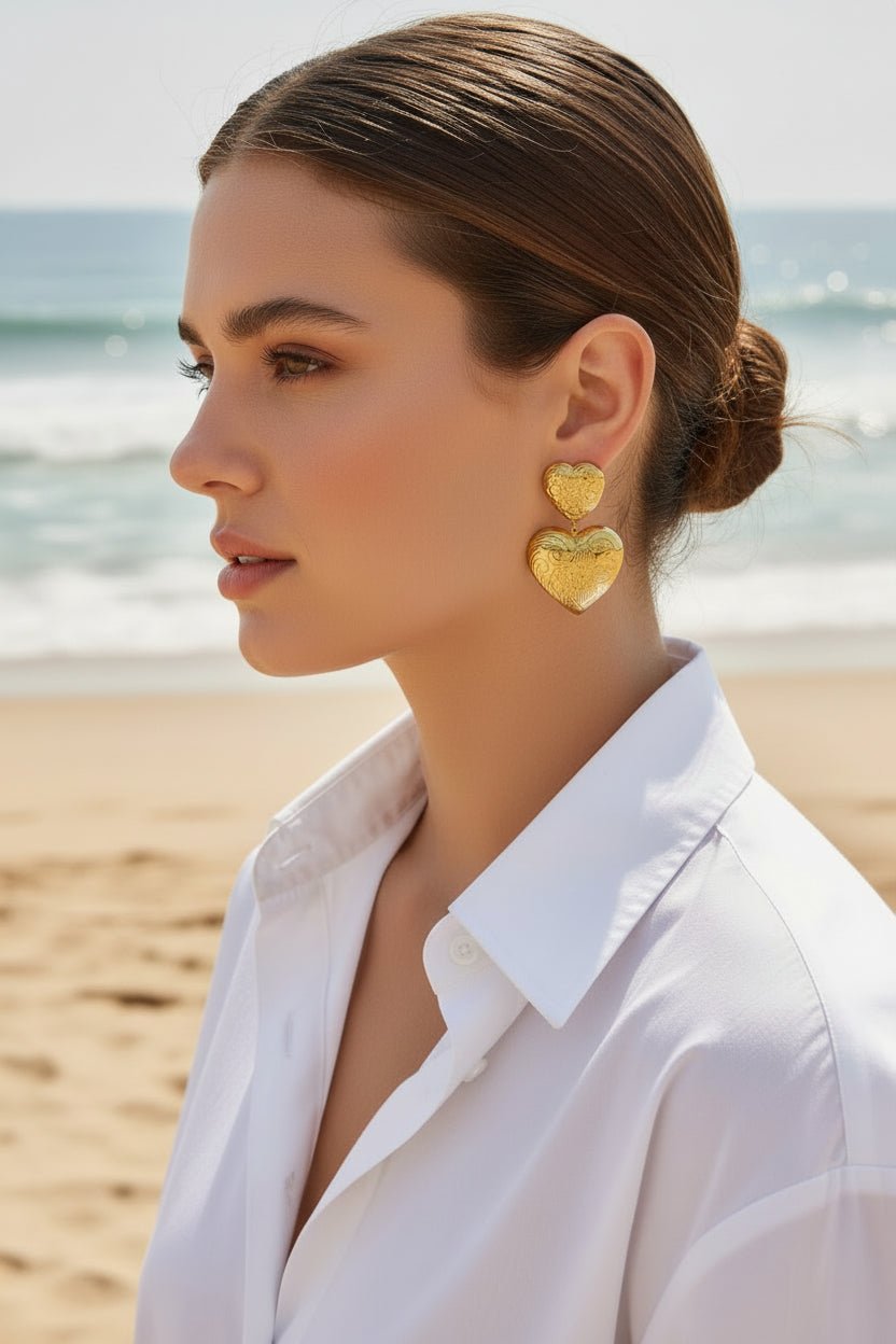 a woman wearing an ornate gold double heart earring and white cotton blouse in an outdoor beach setting 