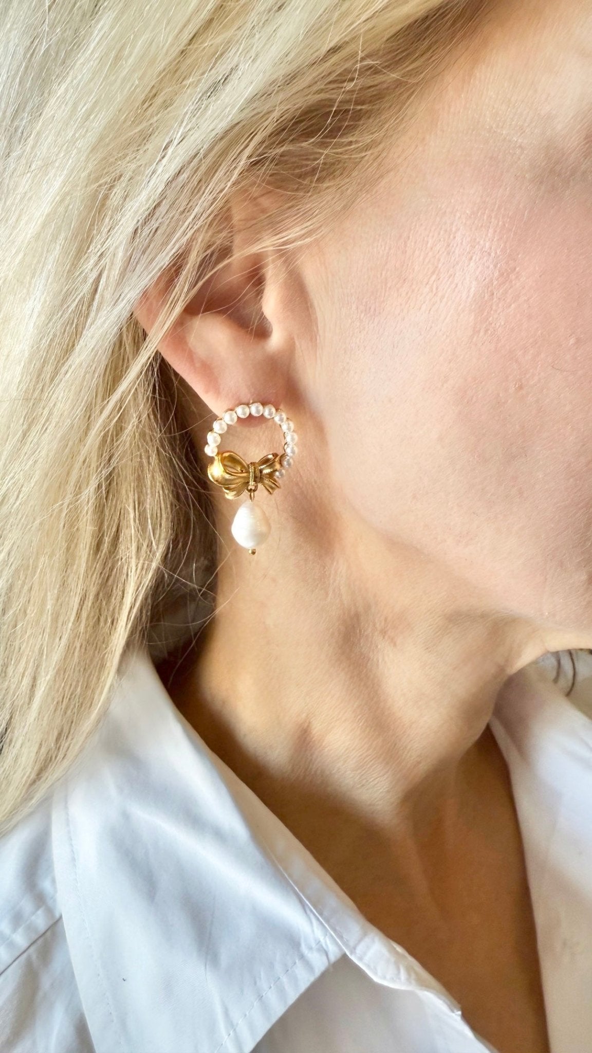 Close-up of a person wearing a gold bow and pearl wreath earring and a white shirt