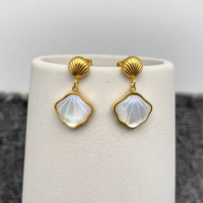 a pair of gold shell charm drop earrings with white resin etched shell design hanging on a white vase