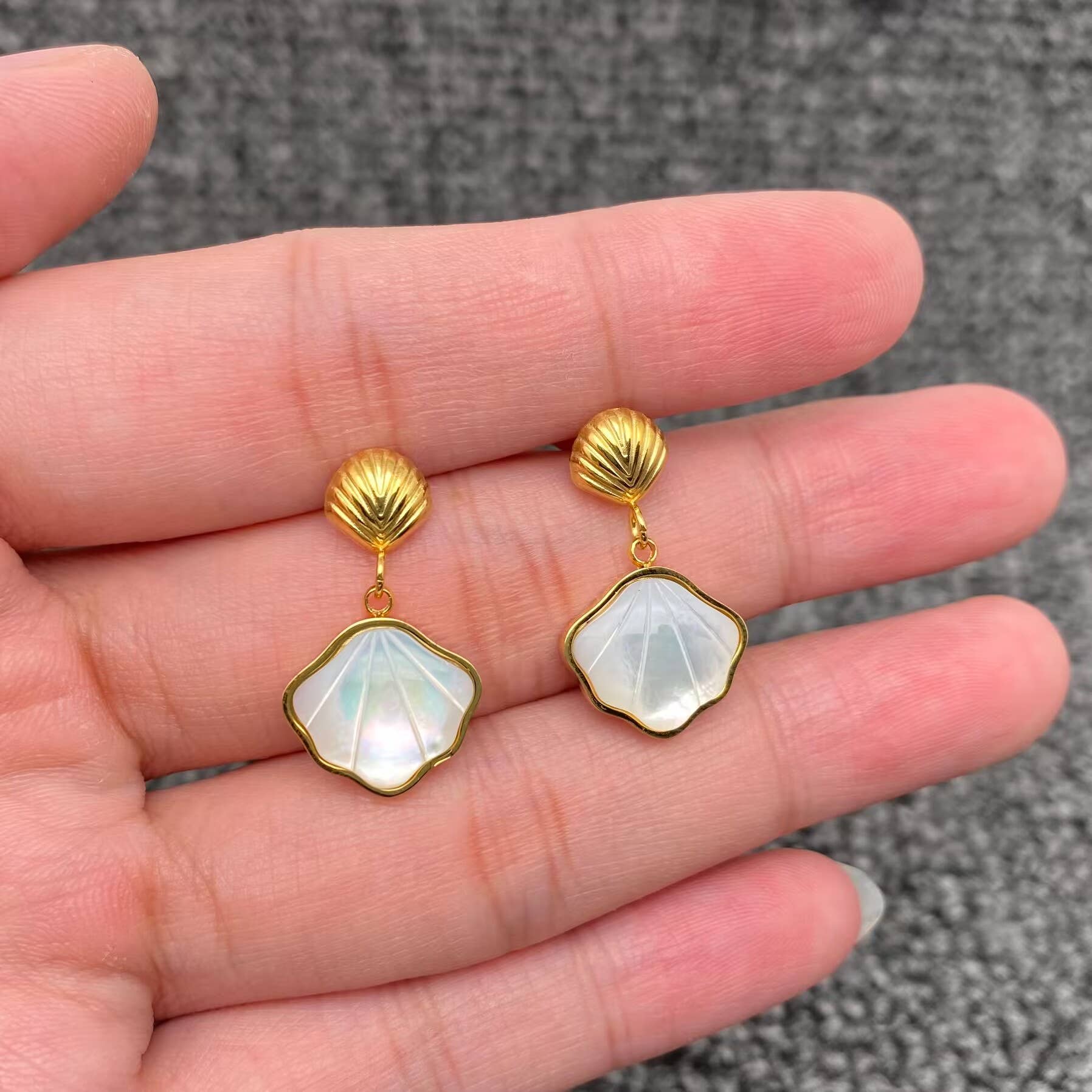 a hand holding a pair of gold shell charm drop earrings with white resin etched shell design