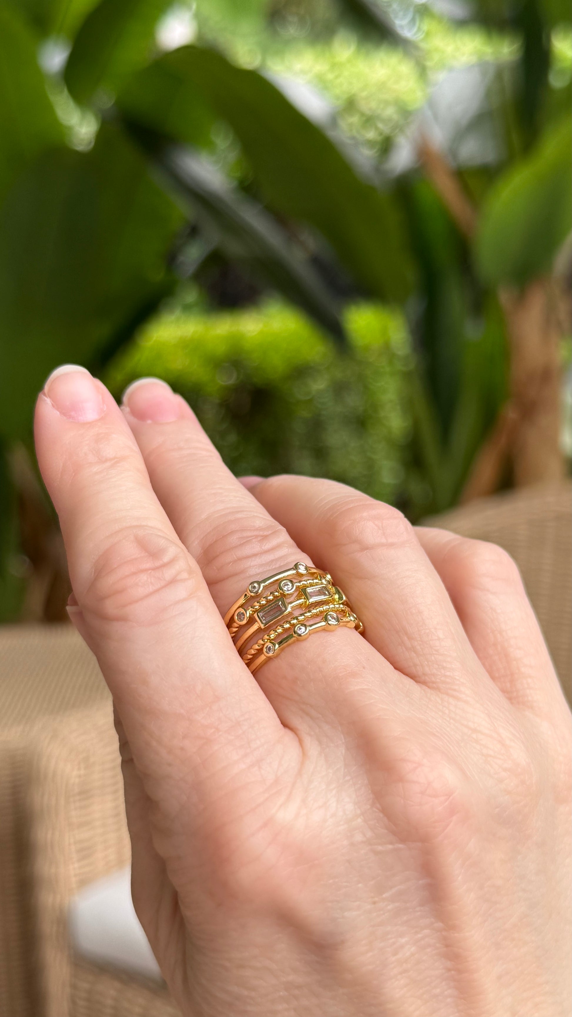Hand wearing a gold stacked ring with crystals with a blurred green outdoor background