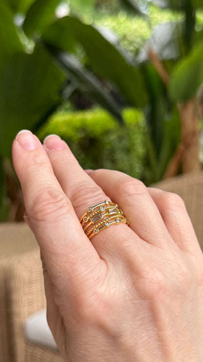 Hand wearing a gold stacked ring with crystals with a blurred green outdoor background