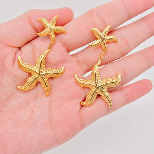 a hand holding a pair of gold starfish shaped dangle earrings