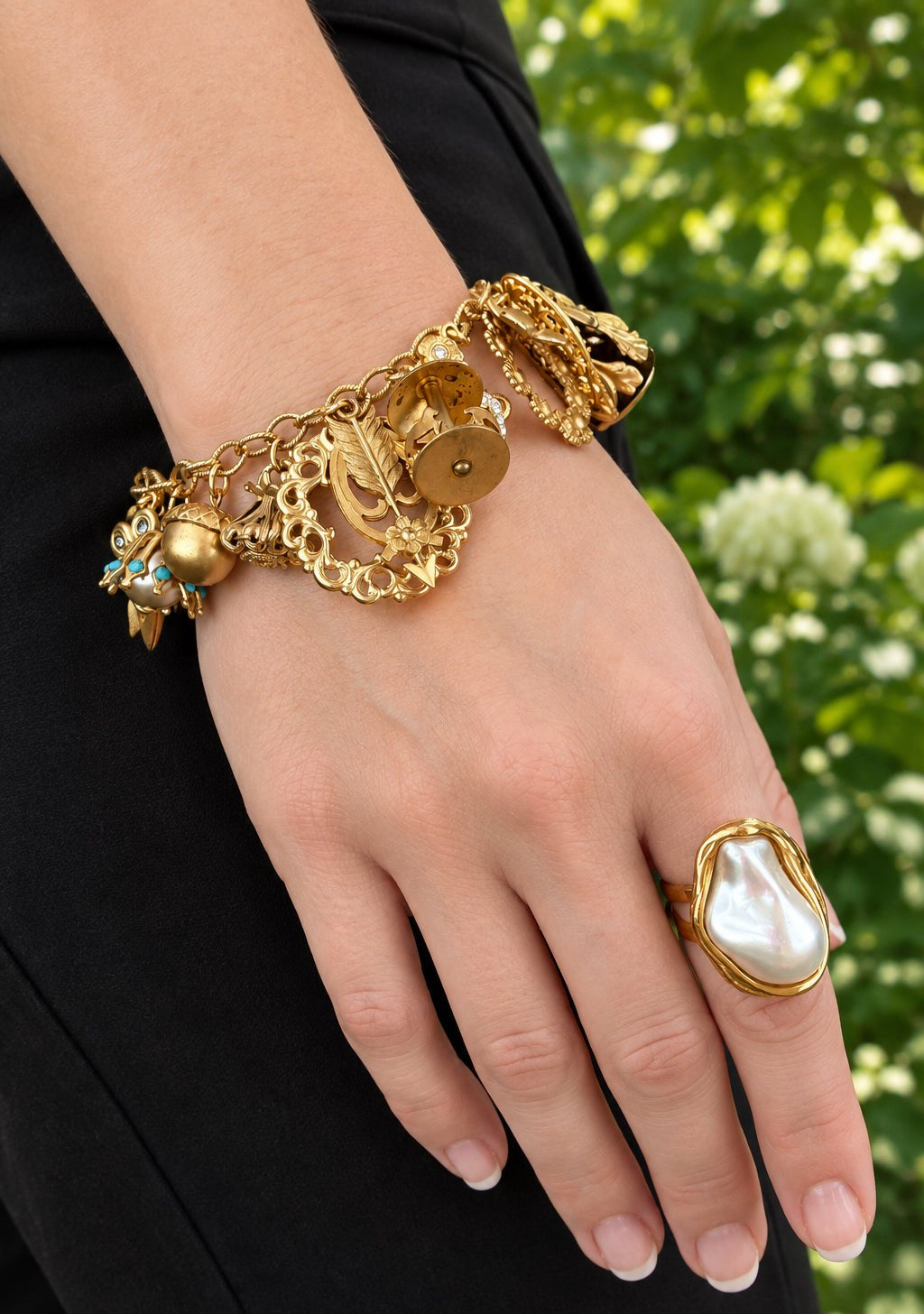 A close-up of a person's hand wearing a chunky gold chain bracelet with charms and a large gold ring with a white pearl, set against black pants outside.