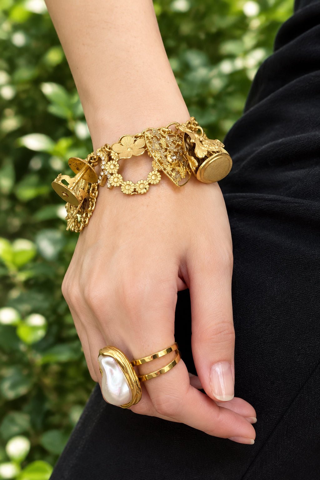 A close-up of a person's hand wearing a gold charm bracelet and gold rings, against black pants in an outdoor setting