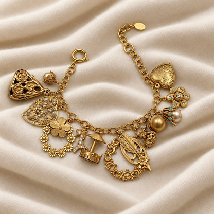 An ornate gold charm bracelet displayed on draped cream-colored fabric. The bracelet features multiple charms, including a heart, a flower with rhinestones, a miniature umbrella, a leaf wreath, and a pearl accent.