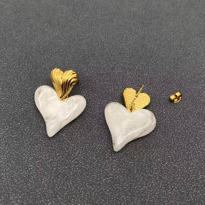 the front and back view of a pair of gold and white resin heart shaped dangle earrings against a grey background