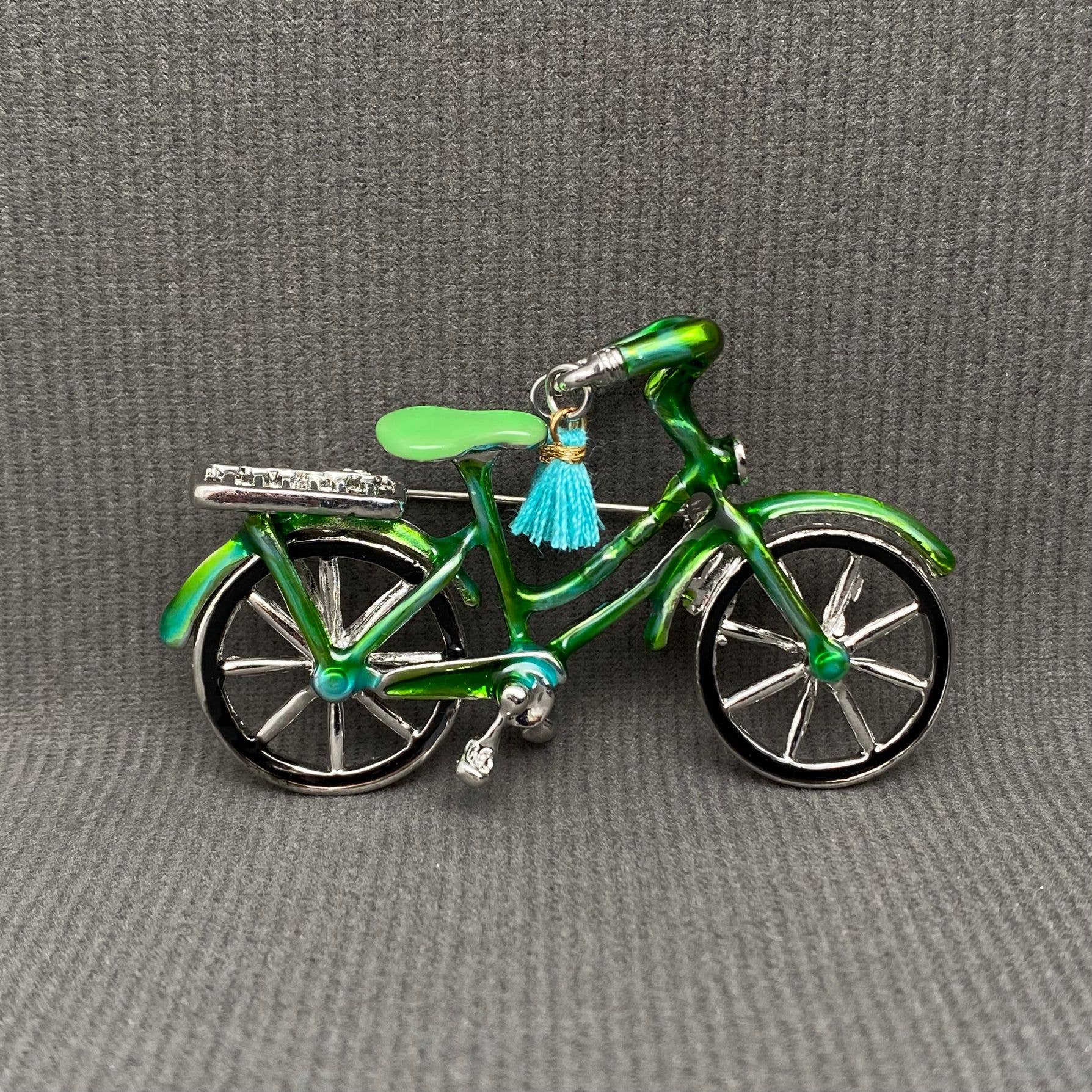 Green bicycle-shaped brooch with a blue tassel on a gray background