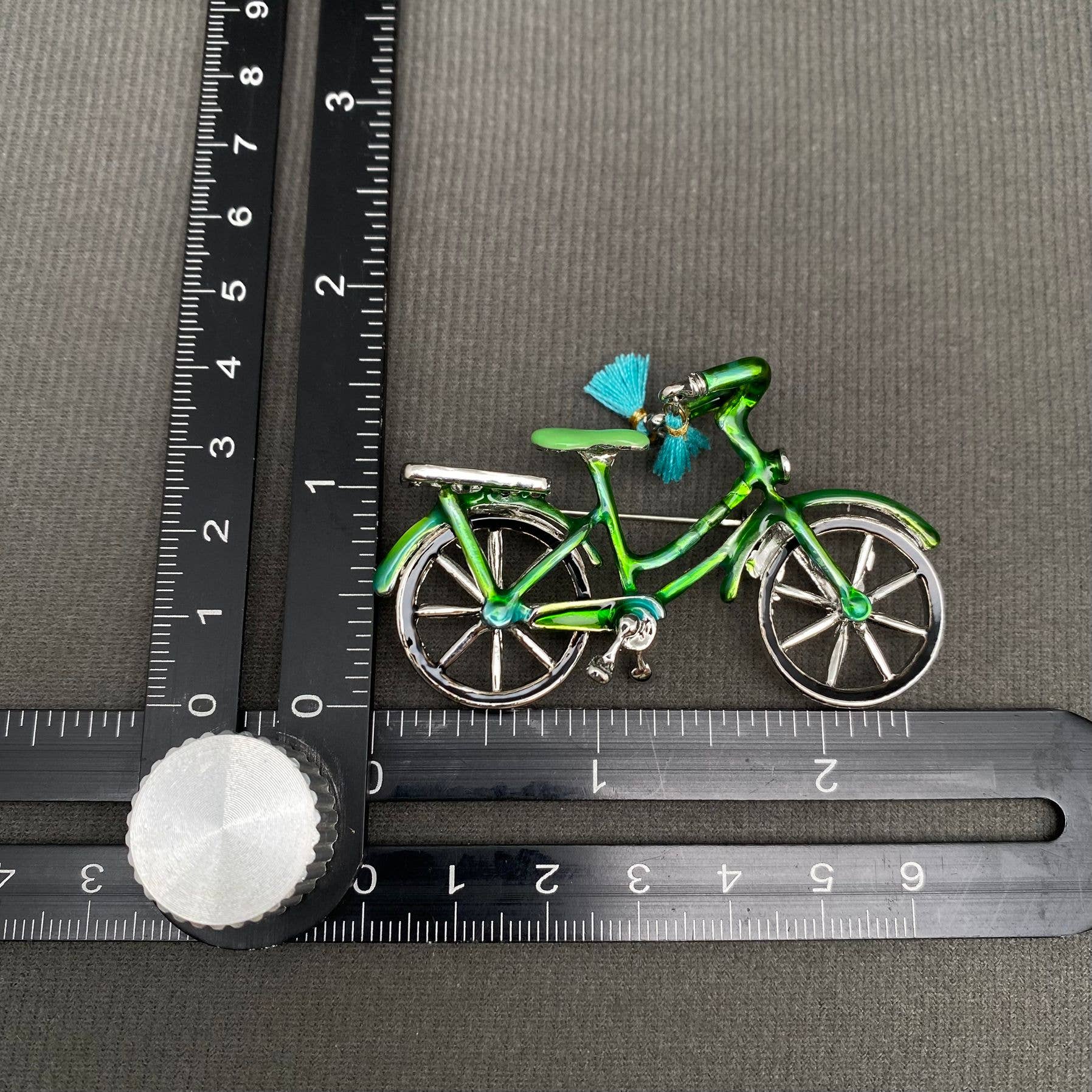 Green bicycle-shaped brooch on a gray surface with a ruler for scale.
