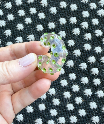 Hand holding a clear acrylic earring with green gemstones against a black and white patterned background