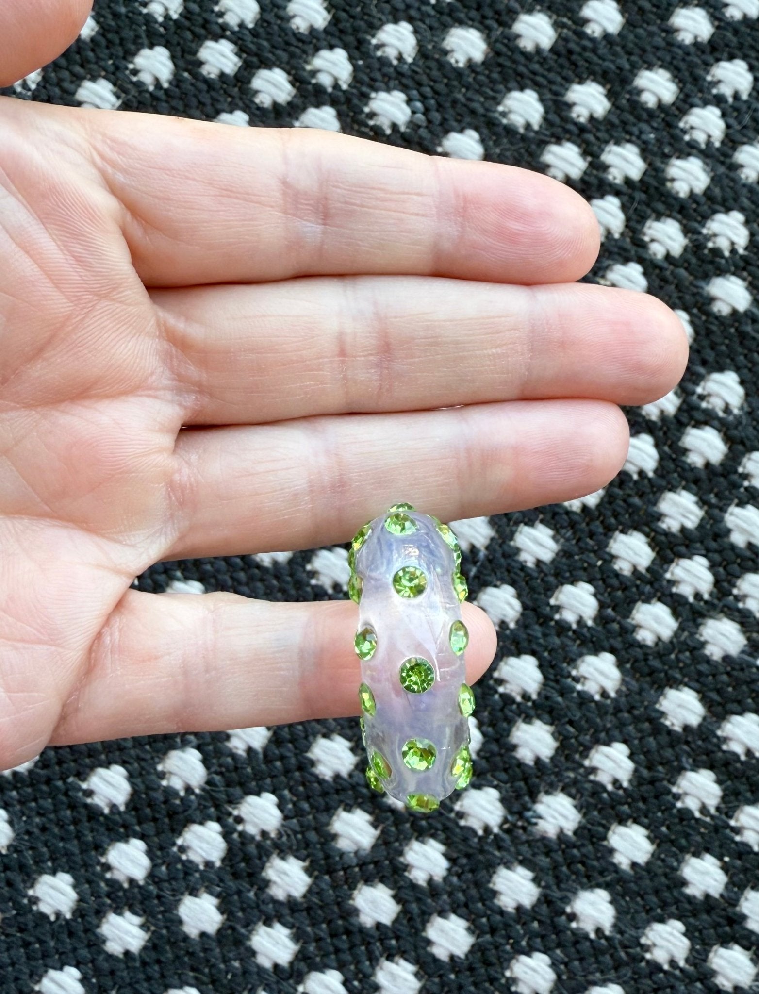 Hand holding a clear acrylic earrings with green rhinestones on a black and white patterned rug background
