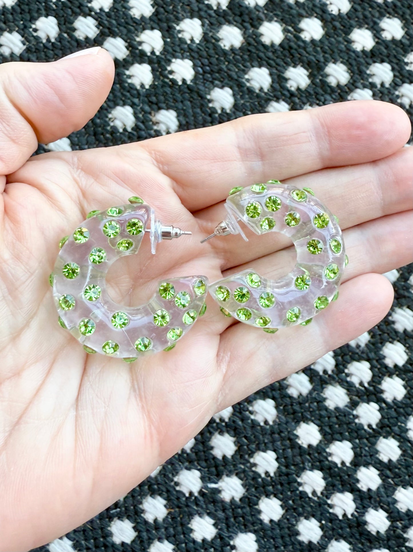 Clear hoop earrings with green gemstones held in a hand against a polka dot rug background