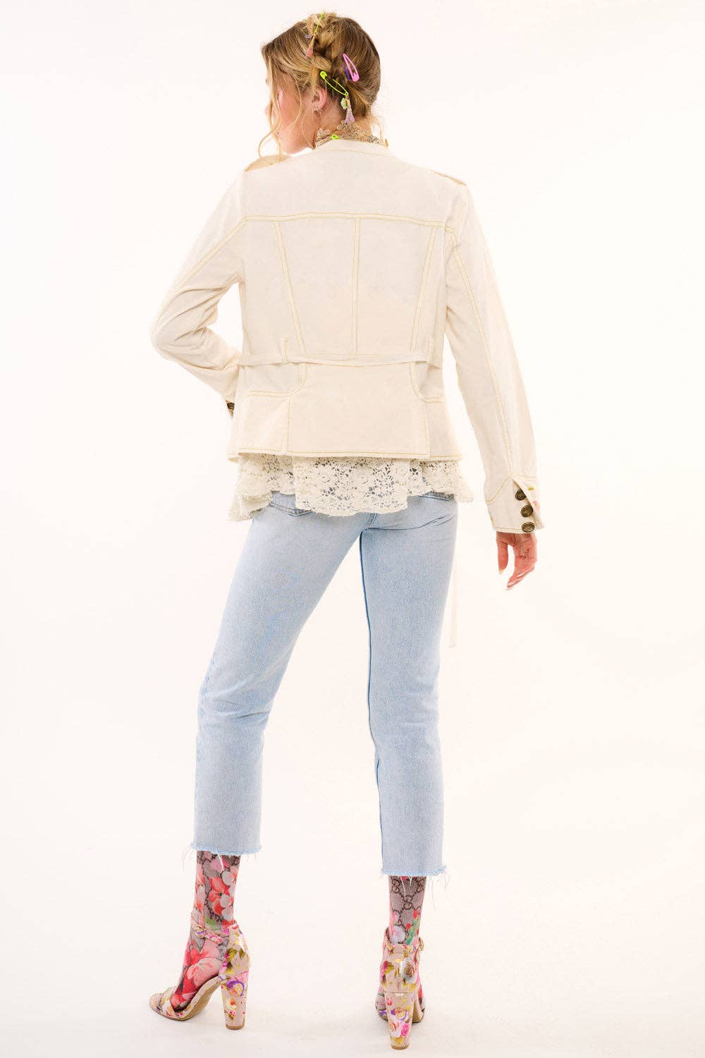 Woman wearing a cream jacket, light blue jeans, and floral shoes on a white background