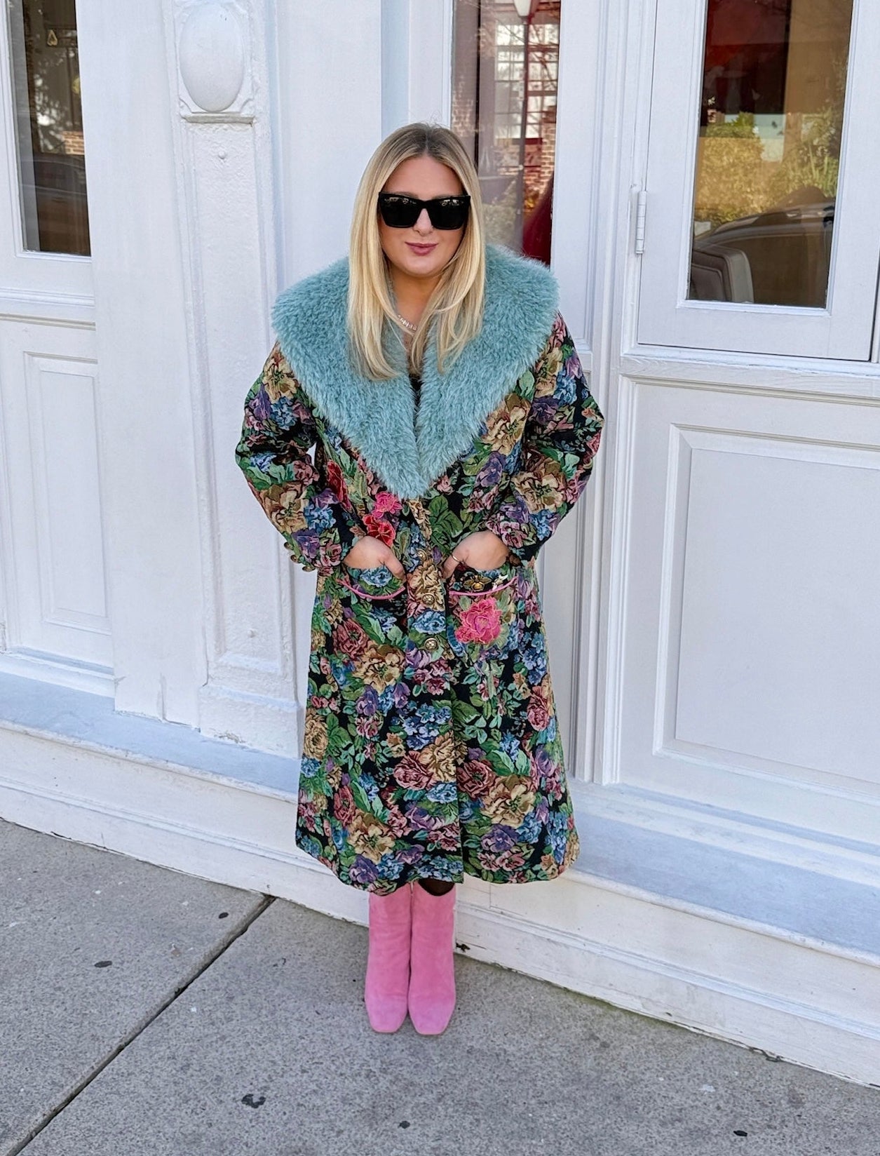 Person wearing a floral coat with a light blue fur collar, pink shoes, and sunglasses standing in front of a white building.