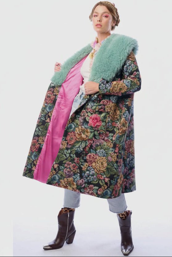 Person wearing a colorful floral coat with a blue fur collar open to show the pink lining on a white background