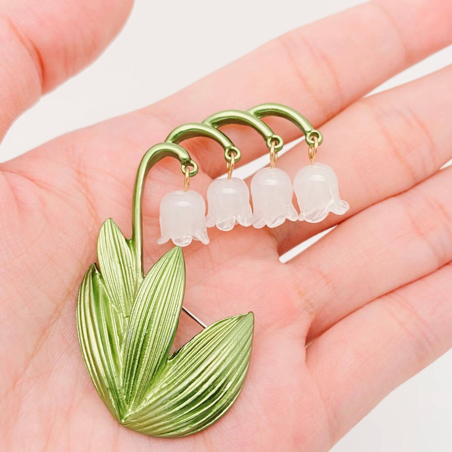 Green and white Lily of the Valley brooch held in a hand against a light background