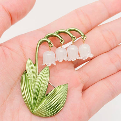 Green and white Lily of the Valley brooch held in a hand against a light background