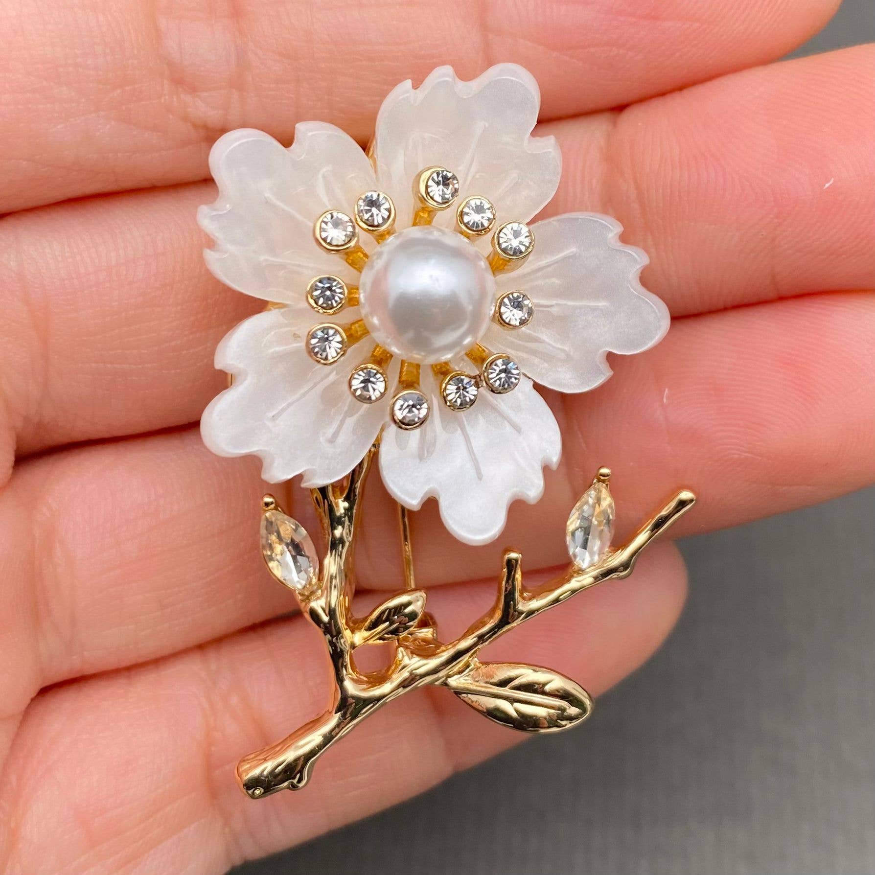 White floral brooch with pearl and crystals held in a hand against a neutral background