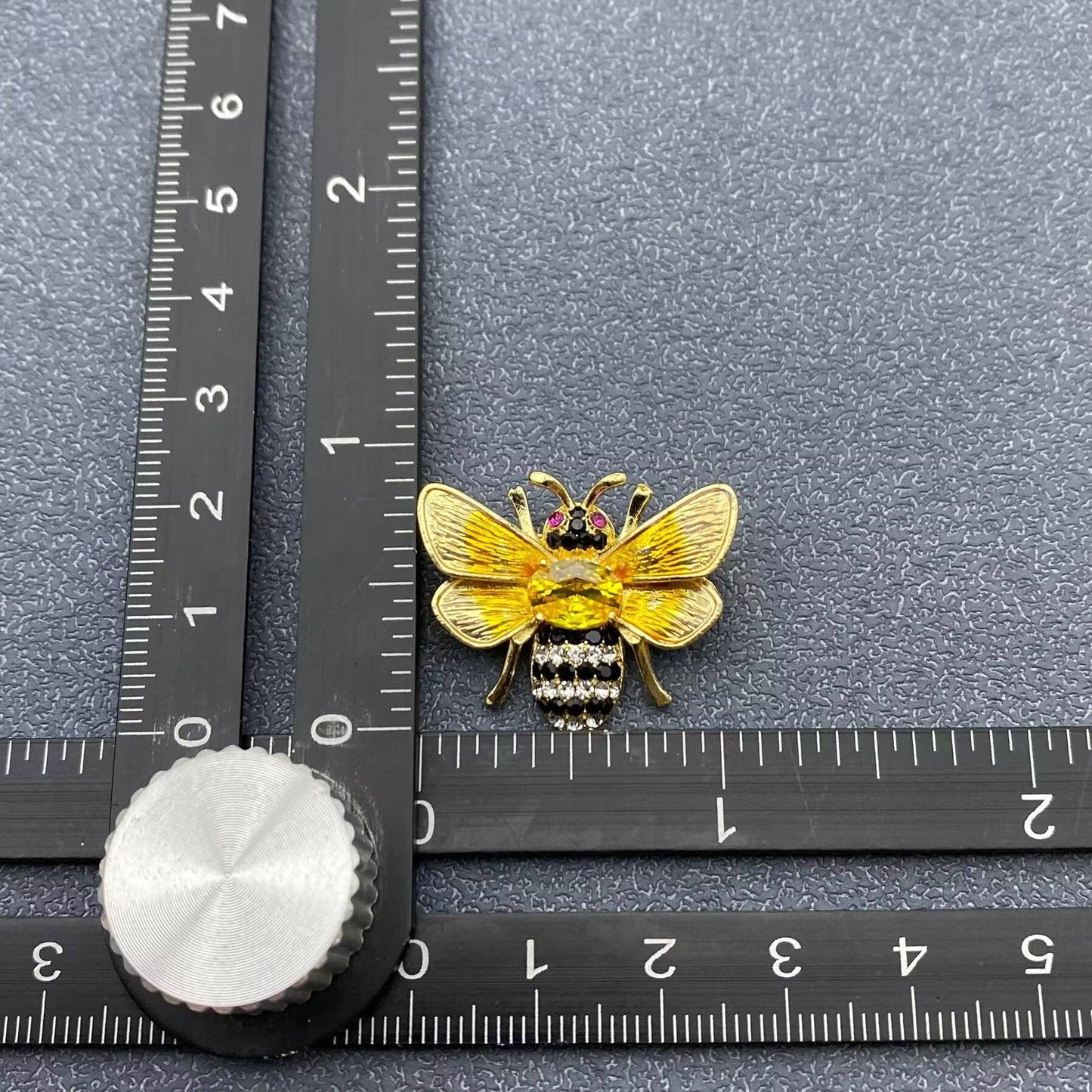Gold bee-shaped brooch on a gray surface with a ruler for scale.