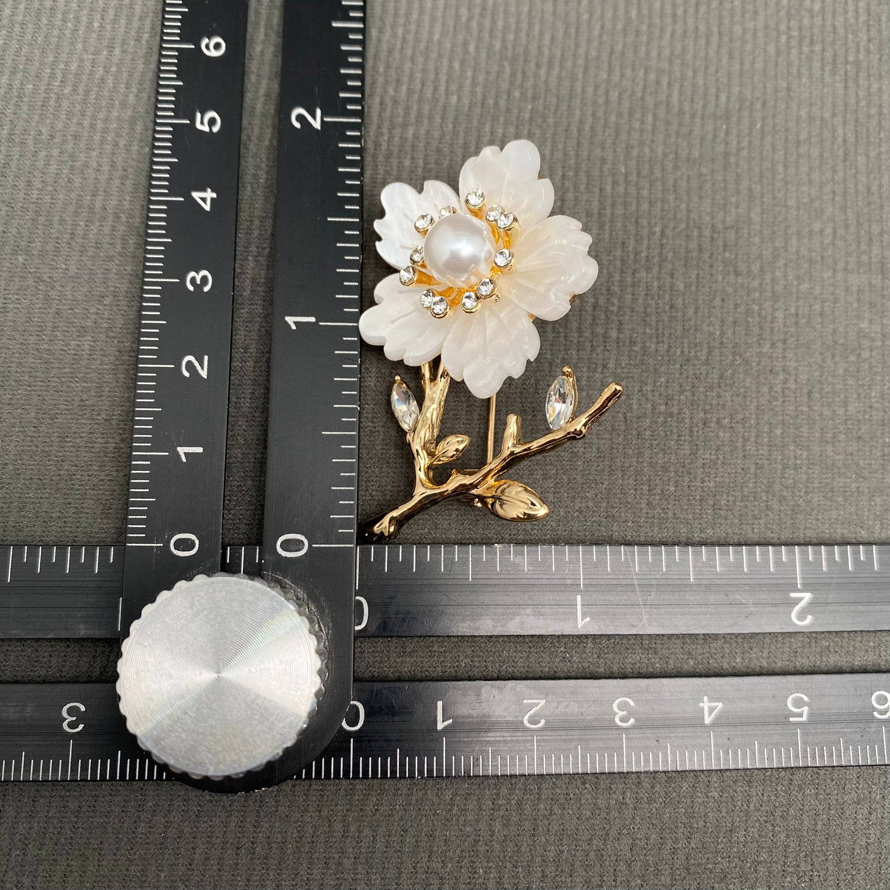 White and gold floral brooch with pearl center on a gray background with rulersfor scale.