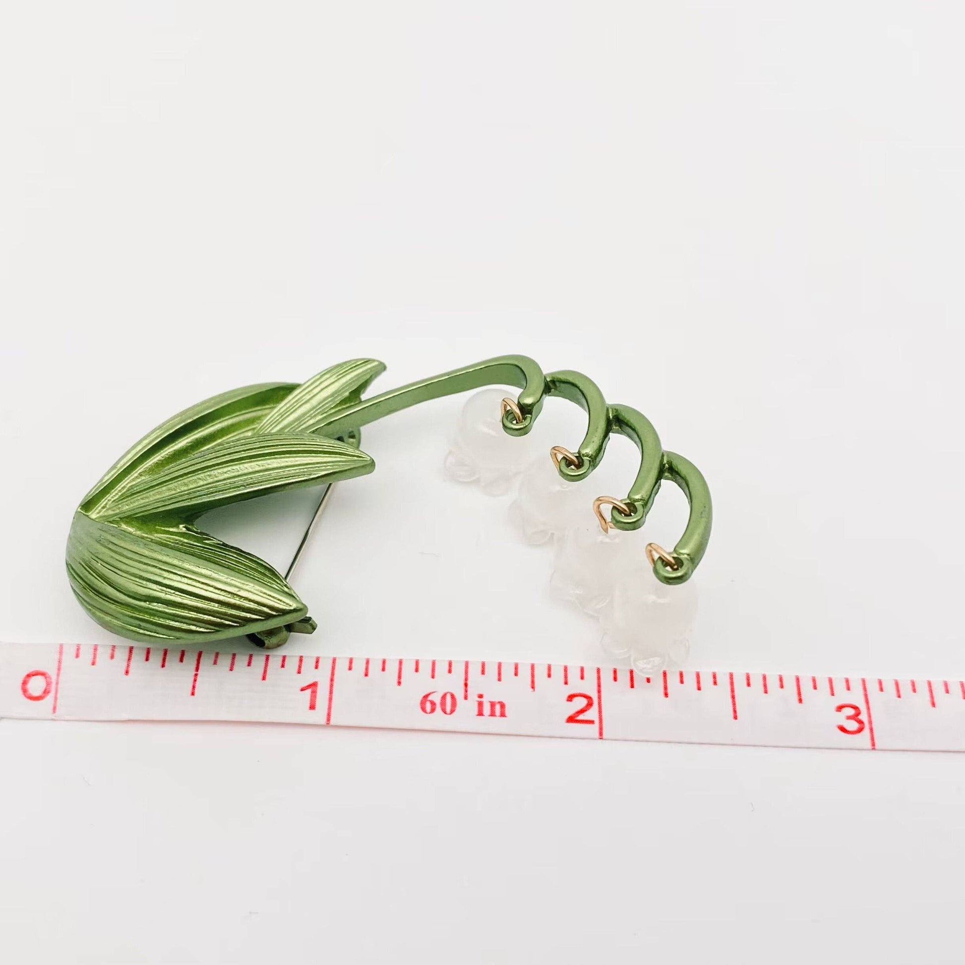 Green and white Lily of the Valley brooch next to a ruler for scale against a white background