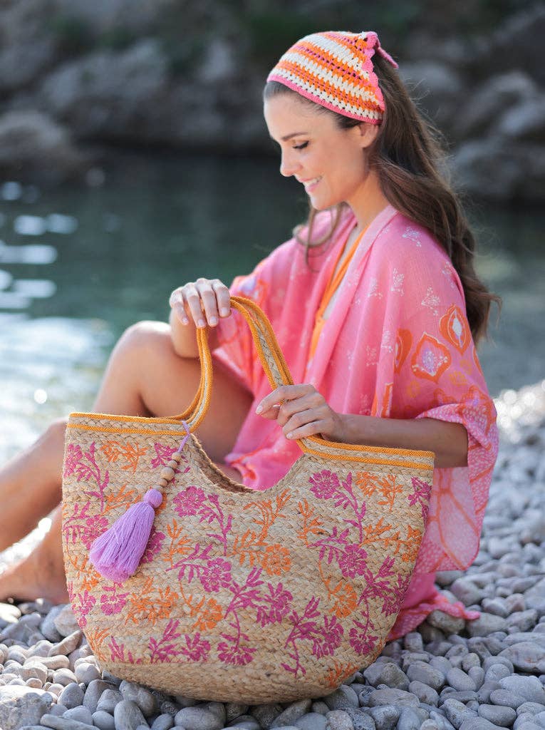 woman sitting on rocks next to a stream with an orange coverup and orange and pink jute tote bag with a purple tassel