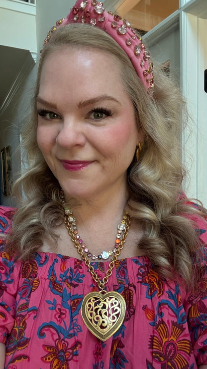 Woman wearing a pink headband and pink floral dress with a gold filigree heart necklace and rainbow colored crystal necklace in an indoor setting