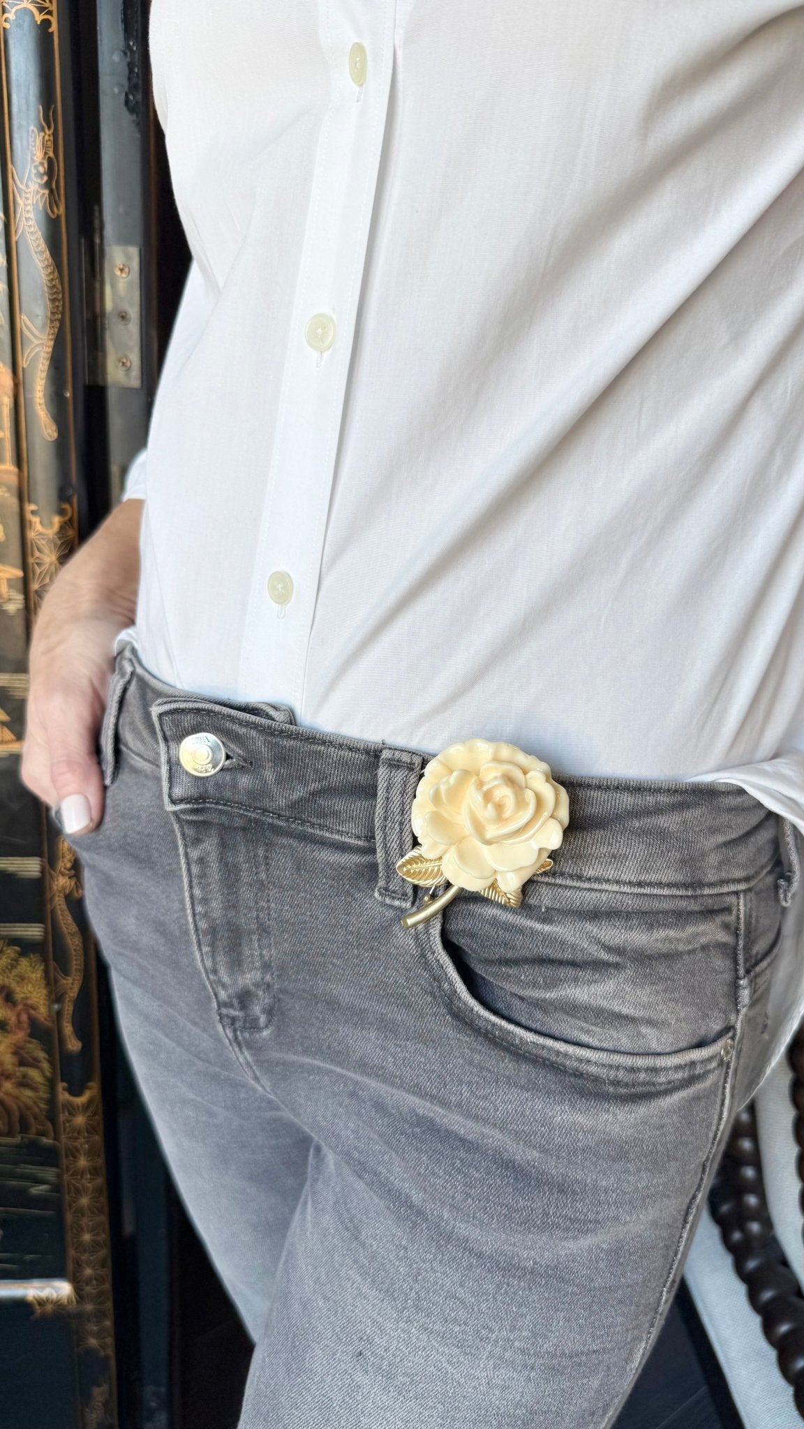 Person wearing a white shirt and gray jeans with a decorative ivory and gold rose brooch on the pocket.